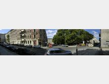 Kreuzung in einer städtischen Wohngegend mit mehrstöckigen Altbauhäusern im historistischen Stil, gepflasterter Straße und parkenden Autos. Auf der rechten Seite stehen große Bäume, daneben gibt es eine Baustelle mit Containern.