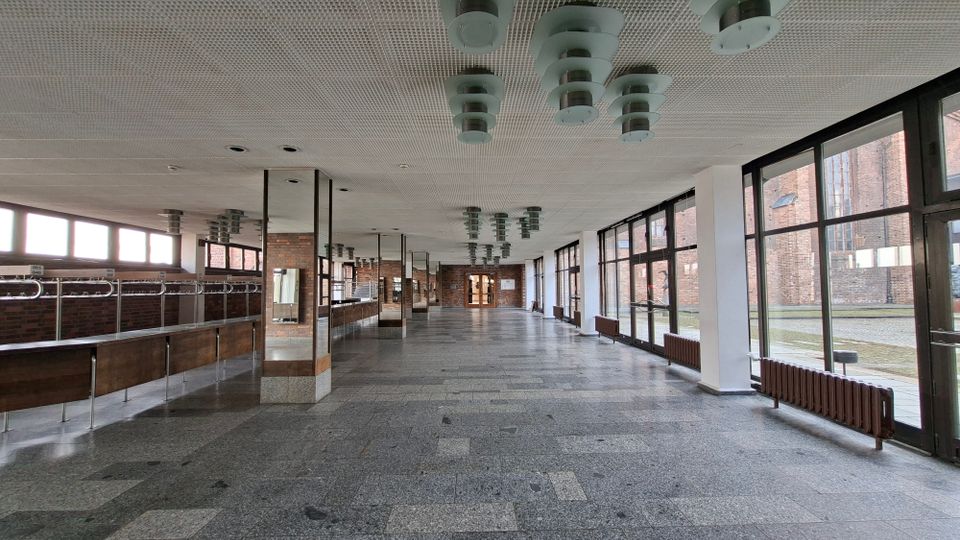 Große, leere Innenhalle mit Steinboden, Rasterdecke und mehreren zylindrischen Deckenleuchten sowie weißen Stützen. Rechts verläuft eine lange Glasfassade mit Blick auf Backsteinbauten und einen Hof; links sind eine Theke bzw. Garderobenanlage und Backsteinwände zu sehen.