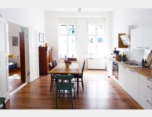 Helle Küche mit Essbereich in einem Altbau mit weiß gestrichenen Wänden, großen Fenstern und Holzdielenboden. Links steht ein Holztisch mit Stühlen, rechts eine Einbauküche mit weißen Fronten und Holzarbeitsplatte; im Hintergrund ein antiker Holzschrank.