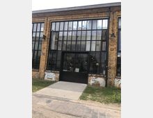 Fabrikgebäude mit Ziegelsteinfassade und großen Fensterfronten, darunter ein schwarzer Eingang mit Glasflügeltüren. Vor dem Gebäude befinden sich ein gepflasterter Weg, etwas Gras und vereinzelt Graffiti an der Wand.