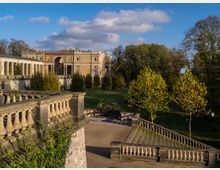 Historische Schlossanlage mit gelber Fassade und Säulenelementen, umgeben von einem weitläufigen Park mit Rasenflächen und Bäumen. Im Vordergrund eine steinerne Balustrade und eine Treppenanlage.