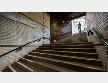 Eine Treppe in einem U-Bahn- oder Bahnhofsbereich, umgeben von gefliesten Wänden. Oben ist ein Ausgang mit Tageslicht und eine Person sichtbar, die die Treppe hinaufgeht; an den Seiten sind Metallgeländer angebracht.