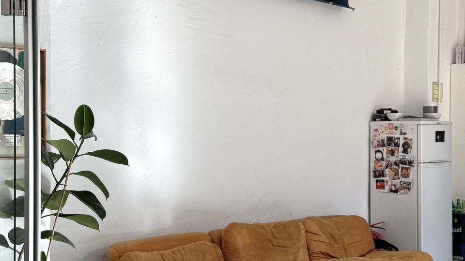 Innenraum eines Büros oder Studios mit weißer Wand, grauem Boden und einer Sitzgruppe aus drei niedrigen braunen Sofaelementen. Daneben stehen eine große Topfpflanze, ein kleiner Kühlschrank mit Magneten sowie über dem Sitzbereich mehrere aufgerollte Hintergrundrollen und ein grünes Notausgangsschild.