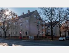 Wohnblock im Stil der Nachkriegsarchitektur mit vier Etagen und Satteldächern, umgeben von kahlen Bäumen. Im Vordergrund verlaufen eine Straße mit Gehwegen, Verkehrsampeln und geparkten Autos.