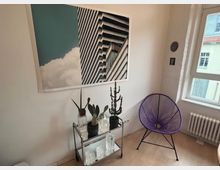 Ein minimalistischer Raum mit hellem Holzboden, einer lila Stuhl, einem Glasregal mit mehreren Dekorationsgegenständen und Pflanzen. An der Wand hängt ein großes gerahmtes Bild mit einem Motiv aus Architektur und Himmel.