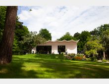 Flaches, modernes Gebäude mit wellenförmigem Dach, großen Fenstern und weißer Fassade, eingebettet in eine grüne Gartenlandschaft mit Rasen, Blumen und Bäumen.