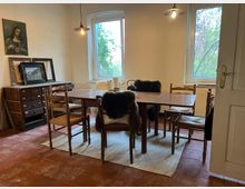 Ein Raum mit einem Holzesstisch und mehreren Stühlen, die mit Fellüberzügen dekoriert sind. Im Hintergrund stehen eine Kommode mit gerahmten Bildern und zwei Fenster, die Blick auf grüne Vegetation bieten; die Wände sind schlicht verputzt und eine Lampe hängt von der Decke.