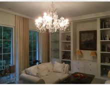 Elegantes Wohnzimmer mit großem Kristalllüster, hellem Sofa und maßgefertigten weißen Einbauregalen mit Vitrinen und Schubladen. Große Fenster mit bodenlangen Vorhängen geben den Blick ins Grüne frei; an der Regalwand hängen ein gerahmtes Bild und stehen zwei Tischlampen.