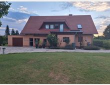 Ein Einfamilienhaus mit orangefarbener Fassade und rotem Satteldach auf einem grasbewachsenen Grundstück, flankiert von einem gepflasterten Hof und Gartenpflanzen. Im Hintergrund sind Bäume und ein leicht bewölkter Himmel zu sehen.