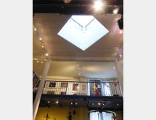 Innenansicht eines mehrstöckigen Museums oder Ausstellungsraums mit einem rechteckigen Oberlicht im Dach, das Tageslicht hereinlässt. Sichtbare Galerieebene mit Geländer, an den Wänden hängen Kunstwerke, dazu punktuelle Deckenbeleuchtung und Säulen im Raum.