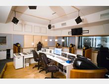 Tonstudio mit modernem Mischpult und mehreren Monitoren, umgeben von großen Lautsprechern und schallisolierten Glasfenstern. Die Wände sind teilweise mit schallabsorbierenden Elementen verkleidet, und die Decke ist mit hängenden Lautsprechern ausgestattet.