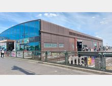 Außenansicht eines modernen Bahnhofsgebäudes mit einer Fassade aus braunen Platten und großen Glasflächen. Im Vordergrund befindet sich eine Metallbrüstung mit Graffiti, neben dem Eingang sind Passanten zu sehen.