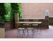 Ein kleiner Außenbereich mit roten Bodenplatten, umgeben von einer niedrigen Backsteinmauer. Im Vordergrund steht ein Set aus Holz- und Metallstühlen mit passendem Holztisch; im Hintergrund wächst grüne Kletterpflanze an Holzpfählen und die Sicht wird von einer Backsteinwand begrenzt.