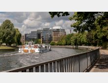 Ein Fluss mit einem Ausflugsschiff im Vordergrund, umgeben von Grünflächen und Bäumen auf der linken Seite und einer Promenade mit Geländer rechts. Im Hintergrund moderne Bürogebäude mit Glasfassaden und bewölktem Himmel.