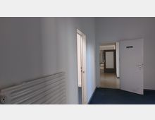 Ein schlichter Innenflur mit weißen Wänden und blauem Teppichboden, einer Heizkörperleiste an der linken Wand und einem offenen Türdurchgang auf der rechten Seite mit der Beschilderung "Ausgang". Im Hintergrund ist eine weitere Tür mit Glasfüllung sichtbar.