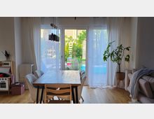 Heller Wohn- und Essbereich mit Holzboden, Esstisch und Stühlen sowie großer Glas-Schiebetür mit weißen Vorhängen zur Terrasse und in den Garten. Rechts stehen eine Topfpflanze und ein Sofa, links ist eine kleine Küchen- bzw. Spielküchenecke zu sehen.