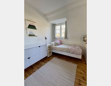 Helles, minimalistisches Schlafzimmer mit weißem Einzelbett unter einem Fenster mit Vorhängen, Holzboden und hellem Teppich. An der Wand stehen eine weiße Kommode und ein kleiner Nachttisch mit Lampe; daneben hängen ein Wandbild und ein kleines Regal.