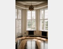 Heller, leerer Raum mit hohen, weißen Sprossenfenstern in einer Erkerfront, durch die Tageslicht auf einen glänzenden Parkettboden mit Muster fällt. An der Decke hängt eine dekorative Pendelleuchte; unter den Fenstern sind schwarze Heizkörper und links eine verglaste Tür.