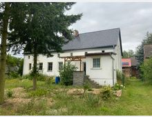Weiße ländliche Hausfassade mit grauem Ziegeldach, kleinen Fenstern und überdachter Eingangstreppe. Im Vordergrund befinden sich ein Garten mit Bäumen, Sträuchern und einer Rasenfläche sowie eine blaue Tonne.