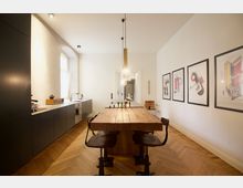 Minimalistisch gestaltete Küche mit einem langen Holztisch in der Mitte, flankiert von schwarzen Stühlen. Auf der linken Seite ist eine matte schwarze Einbauküche mit Waschbecken, Herd und Fenster zu sehen; an der rechten Wand hängen moderne gerahmte Kunstwerke.