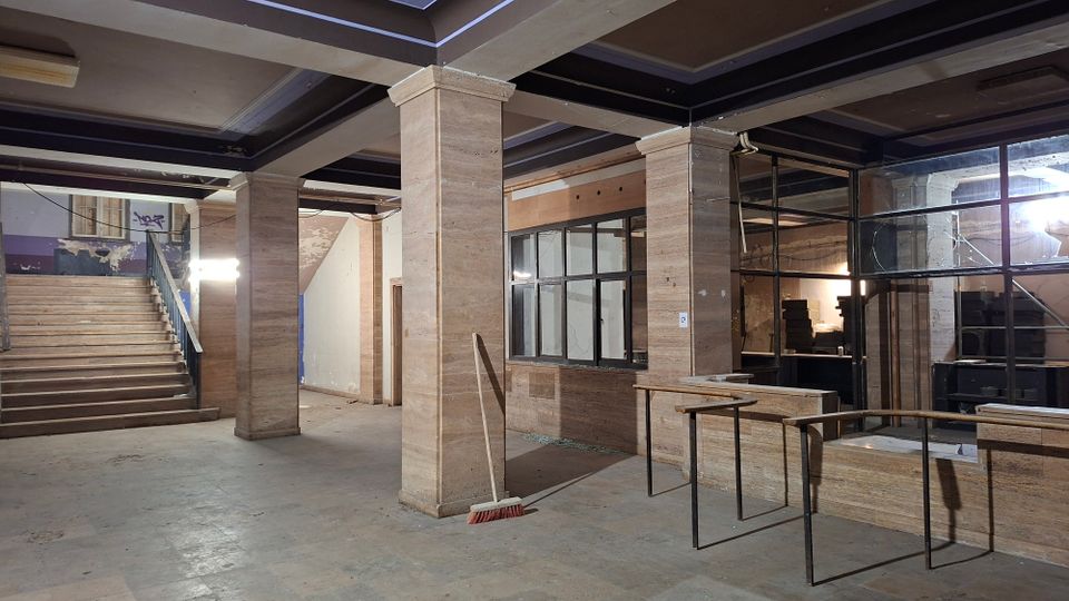 Innenraum einer verlassenen, industriell wirkenden Halle mit Betonboden, massiven Stützen und freiliegenden Deckenbalken. Links führt eine breite Treppe nach oben, rechts sind große Glasfenster und ein niedriger Tresenbereich mit Metallgeländer zu sehen.