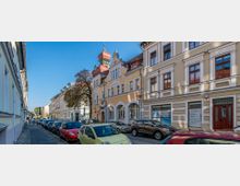 Straßenszene mit historischen Gebäuden, die gelbe und cremefarbene Fassaden mit Verzierungen und roten Dächern aufweisen. Auf beiden Straßenseiten stehen geparkte Autos, und ein Gehweg verläuft entlang der Häuser.