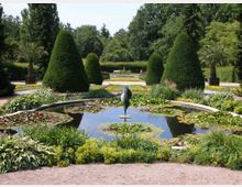 Ein gepflegter Garten mit einem rechteckigen Teich in der Mitte, gefüllt mit Seerosen und umgeben von grüner Vegetation. Der Teich wird von einer modernen Plastikskulptur ergänzt, während kegelförmig geschnittene Büsche und exotische Palmen den Hintergrund bilden.