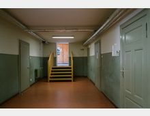 Ein einfacher Flur mit grün gestrichenen Wänden, Türen zu beiden Seiten und einem braunen Boden. Am Ende des Flurs führt eine kleine Holztreppe zu einer hölzernen Tür mit Glasfenster, durch die Tageslicht einfällt.