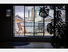 Interior view through large sliding glass doors onto a rooftop balcony with wooden lounge chairs and a railing. Beyond the balcony are brick industrial buildings and several tall smokestacks, with a large leafy plant silhouette in the foreground.