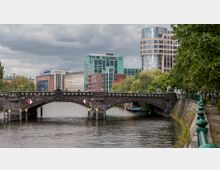 Steinbrücke über einen Fluss, umgeben von modernen und historischen Gebäuden, darunter ein rundes Hochhaus mit Glasfassade und ein rot-brick Gebäude. Am rechten Ufer sind grüne Bäume und ein Gehweg zu sehen, während der Fluss die Umgebung widerspiegelt.