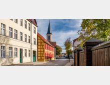 Straßenansicht mit historischen Gebäuden, darunter ein gelbes Fachwerkhaus mit roten Fensterläden und ein beige gestrichenes Mehrfamilienhaus; im Hintergrund ragt der Turm einer Kirche mit einer Spitzhaube in den blauen Himmel. Auf der rechten Seite grenzen ein Holzzaun und Bäume die Straße ein.
