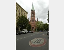 Rote Backsteinkirche mit einem hohen grünen Spitzdach und Uhr über dem Eingangsportal, umgeben von Bäumen und Autos, in einer städtischen Umgebung an einer Straße mit aufgemaltem „30“-Schild auf dem Asphalt. Links ist ein mehrstöckiges Wohngebäude zu sehen.