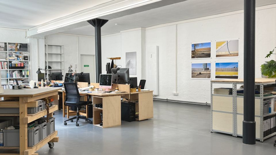 Heller, offener Büroraum mit Betonboden, weißen Wänden und schwarzen Stützsäulen, beleuchtet durch lange Deckenleuchten. Mehrere Holzschreibtische mit Computerarbeitsplätzen, Regale mit Büromaterial sowie an der Wand befestigte Fotodrucke und ein Metallregal prägen den Raum.
