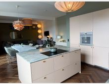 Open-plan kitchen and dining area with a marble-topped island featuring a built-in cooktop, pale cabinetry with an integrated wall oven, and herringbone wood flooring. The space extends into a dining table and lounge area, lit by multiple pendant lights and accented with mirrors and indoor plants.
