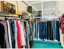 Ein Secondhand-Laden mit bunten Kleidungsstücken, die auf Kleiderstangen hängen, darunter Hosen auf der rechten Seite und Oberteile auf der linken. Im Hintergrund eine weiße Wand mit Retro-Audiogeräten, gelben Pflanzentöpfen und Glasfenstern.