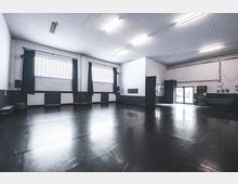 Ein leerer, moderner Tanz- oder Proberaum mit dunklem Bodenbelag, hohen weißen Wänden und großen Fenstern mit Glasbausteinen, die von Vorhängen teilweise verdeckt sind. Die Decke ist mit einem Gitter versehen, und die Beleuchtung erfolgt durch helle Leuchtstoffröhren.