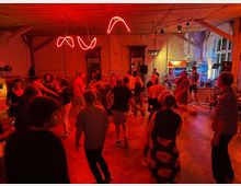 Großer Veranstaltungsraum mit Holzboden und sichtbaren Deckenbalken, beleuchtet von roten Neonröhren und Bühnenscheinwerfern. Rechts steht eine kleine Bühne mit Musikanlage und Instrumenten vor hohen Fenstern und einer teils abgenutzten Wand.