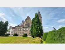 Historisches Steingebäude mit prächtiger Fassade und Turm, umgeben von gepflegten Grünflächen und hohen Hecken, unter einem klaren blauen Himmel.