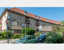 Mehrfamilienhaus im Wohnviertel mit hellbrauner Klinkerfassade, rotem Dach und mehreren Dachgauben sowie Balkonen. Im Vordergrund sind Straßenschilder, geparkte Autos und Büsche zu sehen.