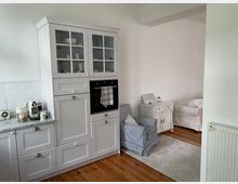 Helle Küchen- und Wohnbereich-Kombination mit weißen Schränken im Landhausstil, einer eingebauten Backofen-Einheit und einer Stein-Arbeitsplatte mit Kaffeemaschine. Im angrenzenden Wohnbereich stehen ein gestreifter Polstersessel, ein kleiner weißer Schrank und ein Teppich auf Holzboden.