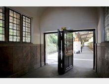 Bahnhofseingang mit geöffneter Holztür, die zu einem Bahnsteig mit gelbem Zug führt; links im Bild befinden sich Glasfenster mit kleinen gerasterten Scheiben, die von dunklem Stein eingerahmt sind.