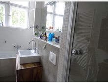 Modernes Badezimmer mit weiß gefliesten Wänden, einem Waschbecken auf einem Holzunterschrank, einer Badewanne mit Wandarmatur sowie einer Glasduschkabine. Im Hintergrund sind Fenster mit Blick ins Grüne zu sehen und zahlreiche Badeutensilien stehen auf einer Ablage vor dem Spiegel.