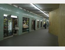 Unterirdischer Fußgängerkorridor mit türkisfarbenen Fliesenwänden, mehreren kleinen Geschäften und Leuchtschriftanzeigen. Über dem breiten Gang sind einfache Decken mit Linienleuchten, rechts ist eine Wand mit Holzplatten verkleidet.