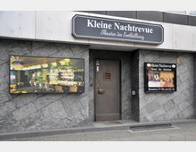 Straßenansicht des Eingangs zur "Kleinen Nachtrevue", einem Theater und Nachtlokal, mit einem schwarz-weißen Schild über der Eingangstür. An der Wand befinden sich links ein beleuchtetes Bild mit einer Bar-Inneneinrichtung und rechts ein Plakat mit Show-Informationen. Die Fassade besteht aus dunklen, diagonal angeordneten Steinplatten.