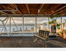 Industrieller Innenraum mit Holzbalkendecke und großen Glasfenstern, die auf ein modernes weißes Wohngebäude mit Innenhof blicken. Im Vordergrund stehen ein schwarzer Designersessel, ein Lautsprecher und mehrere Zimmerpflanzen in Töpfen.