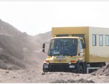 Gelber Lastwagen mit Wohnkabine fährt durch eine sandige, hügelige Wüstenlandschaft. Im Hintergrund sind staubige Erhebungen und die helle, wolkige Atmosphäre der Umgebung sichtbar.