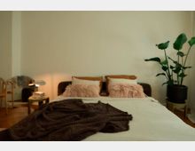 Schlafzimmer mit einem Doppelbett, das mit weißer Bettwäsche, brauner Decke und rosa Kissen dekoriert ist. Links steht ein runder Nachttisch mit einer Lampe, daneben ein Stuhl und ein Spiegel; rechts eine große Zimmerpflanze in einem Topf.