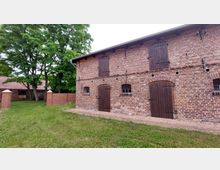 Backsteinbau mit Holztüren und kleinen Fenstern, umgeben von einer Grünfläche und Bäumen; im Hintergrund ein weiteres Gebäude mit Ziegeldach und eine niedrige Backsteinmauer.