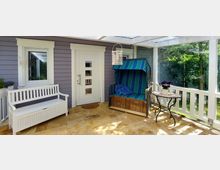 Überdachte Veranda mit Holzfußboden und weißem Geländer vor einer lilafarbenen Hauswand. Links stehen eine weiße Bank und ein Strandkorb mit blau-grün gestreiftem Stoff, daneben ein Tisch mit einer Blume.