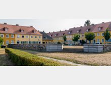 Gelb gestrichene Reihenhäuser mit roten Ziegeldächern stehen um eine offene Grünfläche. Im Vordergrund befinden sich eine niedrige Steinmauer, Treppenstufen und Hecken, während vereinzelte Bäume und parkende Autos die Wohnsiedlung ergänzen.